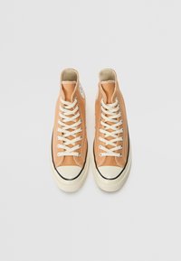 Vysoké plátěné tenisky v hnědo-oranžovém odstínu. Mají krémové tkaničky, bílý špičkový kryt a černé detaily podél podrážky. Viditelná jednoduchá nášivka s logem.