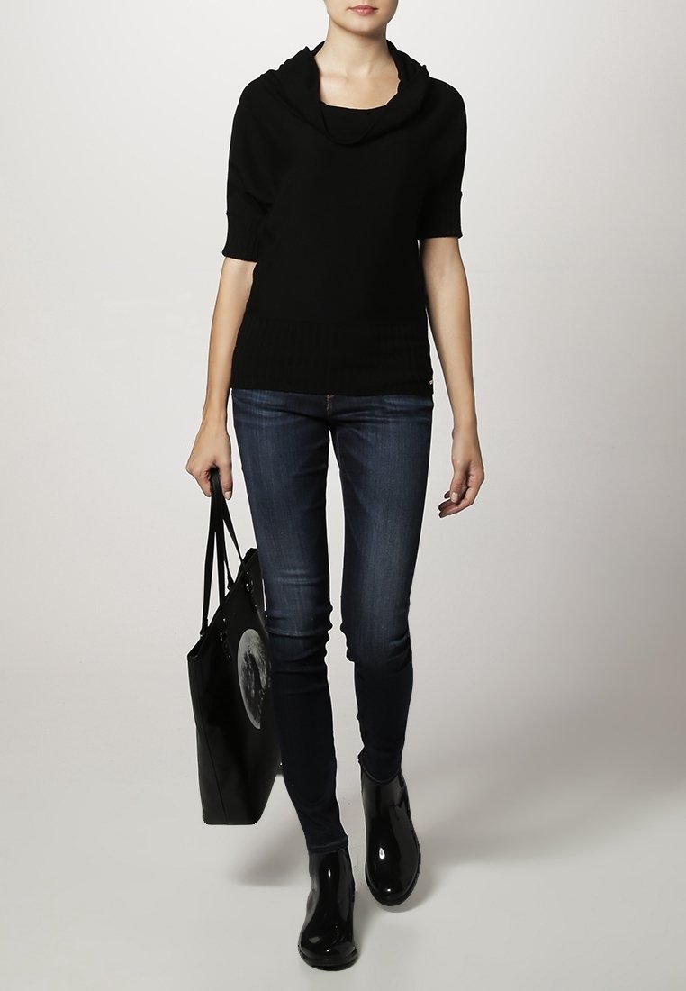 Femme portant un haut noir à col bénitier, un jean skinny foncé et des bottines noires, portant un sac cabas noir avec un motif de lune.