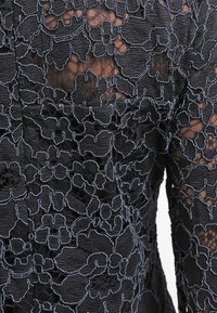 Černá krajka s propracovanými květinovými vzory, obsahující průsvitné části a texturovaný povrch. Modré prošívání zvýrazňuje detaily designu.