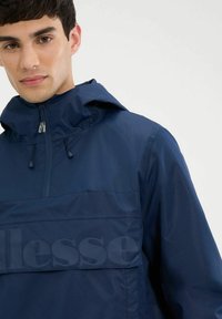 Dunkelblauer Windbreaker aus leichtem Stoff mit einem Reißverschluss am Hals, elastischen Bündchen und einer großen Logo-Panel auf der Vorderseite. Glatte Textur.
