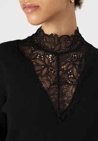 Haut en tricot noir avec un col en dentelle haute ornée de motifs floraux et de bords festonnés. Conception près du corps avec des détails en dentelle raffinés.