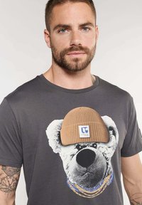Camiseta gris con un gráfico de un oso que lleva un gorro de punto beige en la cabeza, detallada con acentos azules y un logotipo. Manga corta y cuello redondo.