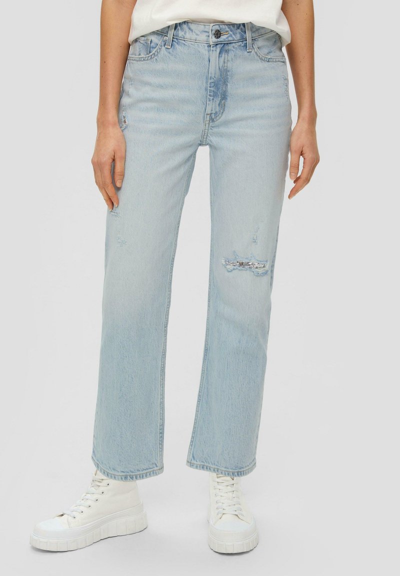 Jeans dritti strappati azzurro chiaro con vita alta e dettagli di scolorimento sottili. Abbinati a sneakers bianche con piattaforma.