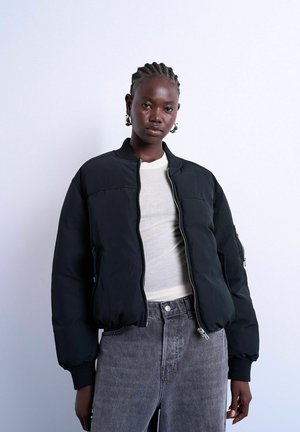 Kvinde med fletninger iført grønne øreringe, en sort dunjakke over en hvid top og højttaljede grå jeans, stående mod en hvid baggrund.