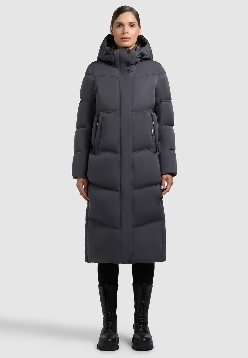 Prošívaná bunda v tmavě šedé barvě s kapucí, kulatým prošíváním, dvěma předními kapsami a dlouhým střihem. Kombinováno s černými botami.
