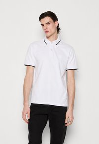 Weißes Poloshirt mit schwarzem Kragen und Ärmelakzenten, kurzenÄrmeln, zwei-Knopf-Leiste und dezentem Logo auf der linken Brust.
