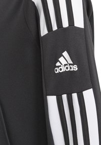 Chaqueta deportiva negra con tejido texturizado, que presenta tres rayas blancas en el hombro y un logo de Adidas bordado en blanco.