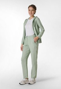 Lichtgroene, lichte jas met capuchon over een witte henley-shirt. Bijpassende lichtgroene, met koord verstelbare broek. Crème en grijze sneakers.