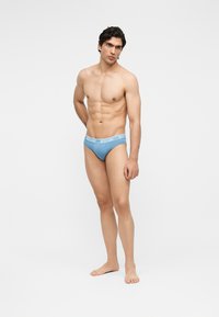 Izmos férfi áll mezítláb, világoskék Moschino alsónadrágot visel, egyszínű fehér háttér előtt, kissé oldalra nézve.