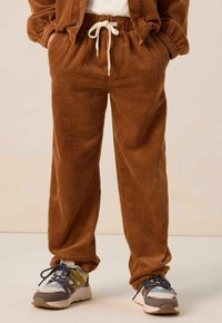 Pantalons en corduroy marron avec une texture en côtes, dotés d'une taille élastique avec un cordon blanc, associés à des baskets sportives multicolores.
