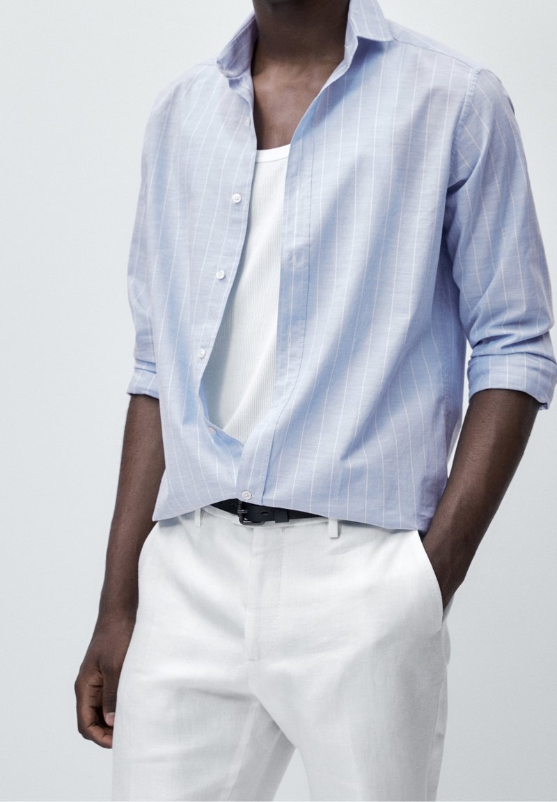 Homme portant une chemise à boutons rayée bleu clair sur un débardeur blanc avec un pantalon blanc, une main dans la poche sur un fond uni.