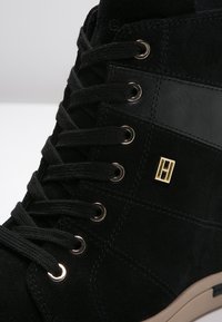 Svart mocka ankelsko med runda snören, metallöglor och en guldsymbol. Har en texturerad sidopanel och en gummisula.