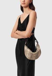 Vaalea beigemallinen python-kuvioinen käsilaukku, jossa on kaareva muoto ja musta olkahihna, jota mallilla on käsissään mustan satiinimekon kanssa.