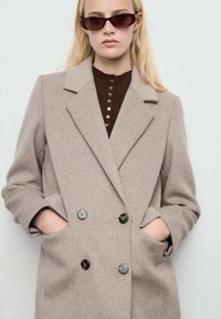 Manteau beige en mélange de laine à double boutonnage avec larges revers, deux poches avant et boutons noirs ; porté par-dessus un haut marron foncé.