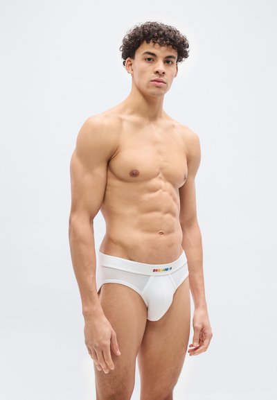 Νεαρός άνδρας με κυματιστά μαλλιά που φοράει λευκά εσώρουχα με πολύχρωμο λογότυπο "DSQUARED2" στη μέση, στέκεται μπροστά σε ένα απλό φωτεινό φόντο.