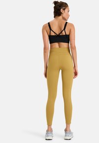 Soutien-gorge de sport noir avec des bretelles croisées, associé à un legging jaune moutarde taille haute. Le mannequin porte des chaussures de sport grises. Tissu lisse et extensible.