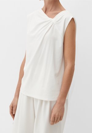 Kobieta w białej bluzce bez rękawów z marszczeniem przy dekolcie, w połączeniu z luźnymi białymi spodniami na jednolitym tle.