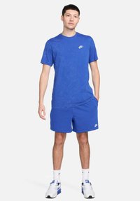 Blå bomulls-T-shirt med korta ärmar och rund halsringning, matchad med blå shorts. Båda har en liten vit Nike-logotyp. Vita sportskor.