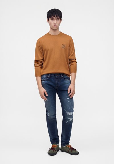 Jeune homme debout portant une chemise marron à manches longues, un jean bleu délavé et des baskets vertes avec des lacets orange sur fond blanc uni.