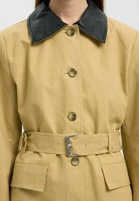 Trench coat khaki con un collo in pelle nera, cinque bottoni frontali, vita con cintura e dettagli con tasche laterali. Il tessuto appare liscio e strutturato.