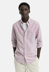 Chemise à rayures rose et bleue. Possède un col, des manches longues, une poche poitrine et est fabriquée en tissu léger.