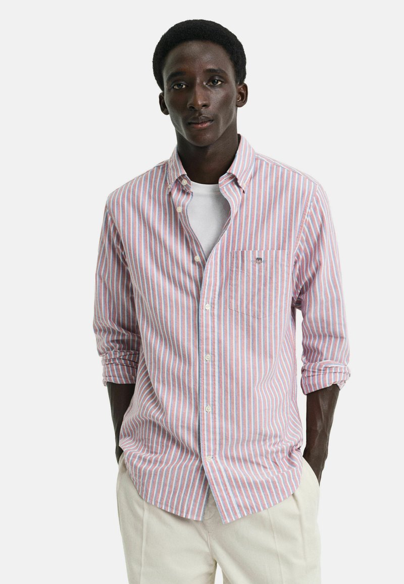 Chemise à rayures rose et bleue. Possède un col, des manches longues, une poche poitrine et est fabriquée en tissu léger.