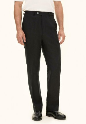 Mężczyzna ubrany w czarne formalne spodnie, czarne skórzane mokasyny oraz białą koszulę z krótkim rękawem, stojący na jednolitym tle.