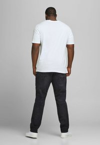 Weißes T-Shirt mit kurzen Ärmeln und dunklen Jeans, von hinten betrachtet. Das T-Shirt hat einen runden Halsausschnitt; die Jeans haben Gesäßtaschen und eine schmale Passform.