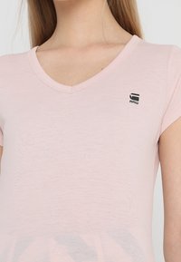 Žena nosí světle růžové tričko s výstřihem do V a malým černým logem "G RAW" na levé horní části hrudi.