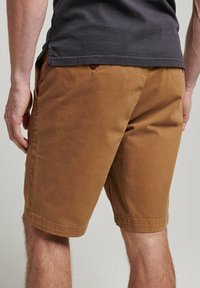 Pantalones cortos de algodón marrón con corte recto, que cuentan con bolsillos laterales y un botón en la parte trasera. Textura suave y diseño hasta la rodilla.