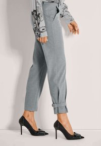 Vrouw die loopt in grijze taps toelopende broek met enkelgespen en zwarte hoge hakken met strikaccenten.