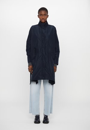 Marineblauer wasserdichter Mantel mit hohem Kragen, zwei Fronttaschen und lässiger Passform, kombiniert mit hellblauen weiter geschnittenen Jeans und schwarzen, polierten Stiefeln.