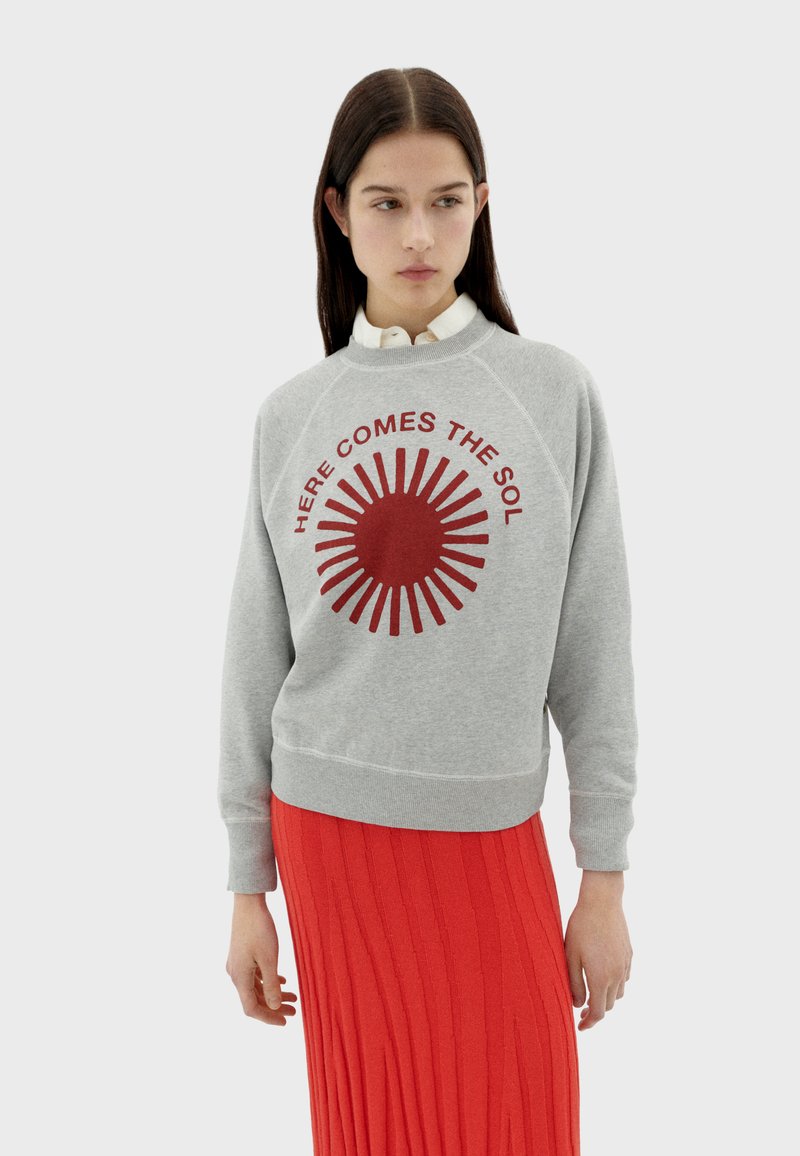Sweatshirt gris avec un graphique de soleil rouge et le texte "Ici vient le SOL," associé à une jupe rouge côtelée. Design décontracté, texture douce.