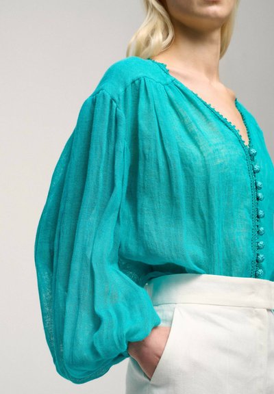 Bluza din in de culoare teal, cu decolteu în V, având nasturi texturați, mâneci puff și un tiv delicat cu dantelă. Purtați cu pantaloni albi cu talie înaltă.