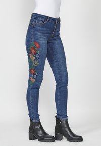 Skinny blue jeans with embroidered floral design in red, blue, and green on the left leg. Features five pockets and a mid-rise waistband.