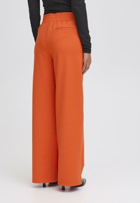 Oranje wijde broek met elastische tailleband en achterzak, gemaakt van zachte stof, gecombineerd met zwarte hoge hakken.
