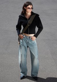 Schwarzer gerippter Reißverschluss-Pullover, hellblaue weit geschnittene Jeans und schwarze spitze Absatzschuhe. Die Accessoires umfassen eine Umhängetasche mit Hardware.