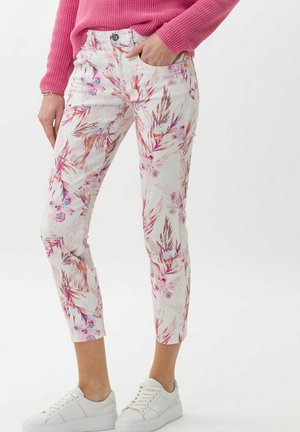 Kvinde iført hvide cropped bukser med pink og lilla blomsterprint, lyserød strikket sweater og hvide sneakers, stående med den ene hånd i lommen.