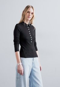 Camisa polo de manga larga negra con una tela texturizada, que presenta una abertura con botones y un diseño ajustado. Combinada con unos pantalones vaqueros de pierna ancha en azul claro.