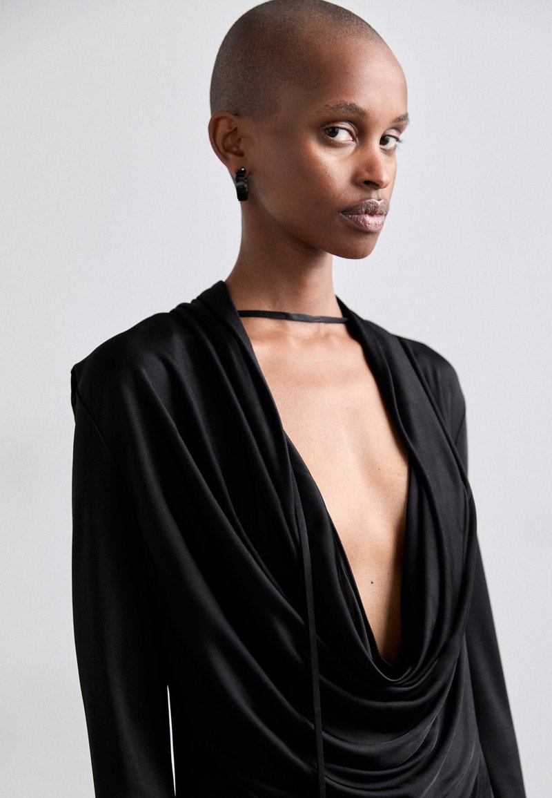 Black draped dress with deep V-neck, long sleeves, and a thin neck tie. Smooth fabric, subtle sheen, and a layered design detail.