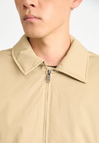 Close-up of a person wearing a beige zip-up jacket with a collar, and a small hoop earring in the left ear.