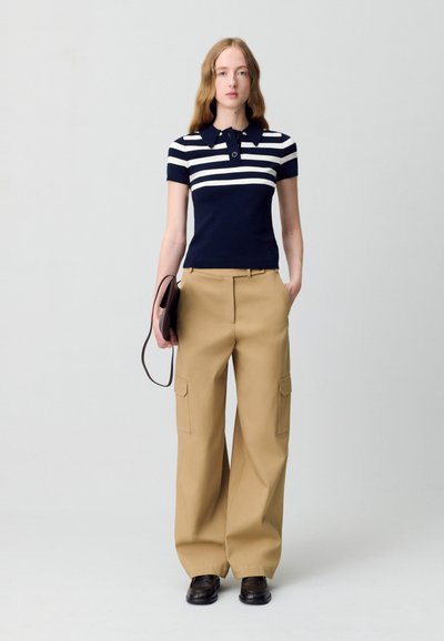 Polo a righe blu navy e bianche con maniche corte, abbinato a pantaloni beige a gamba larga con tasche cargo. Il modello tiene una pochette scura.