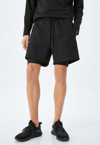 Sorte træningsshorts med elastisk talje og snøre, der har en løs pasform og sidelommer, parret med sorte sportssko.