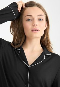 Camisón negro con ribete blanco en el cuello y la tapeta de botones, que presenta una textura suave y lisa, con un ajuste holgado.