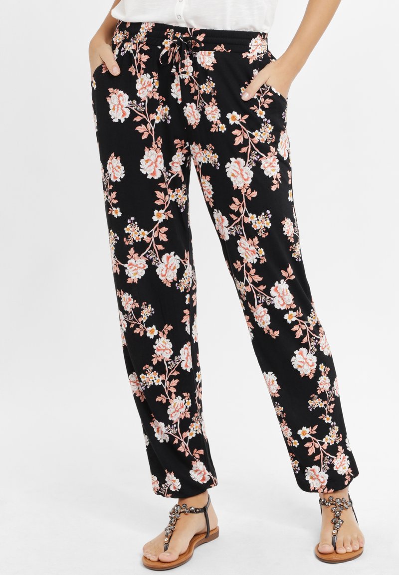 Pantaloni neri con un motivo floreale in rosa e bianco, dotati di una cintura elastica e tasche, abbinati a sandali con flip-flop adornati di gioielli.