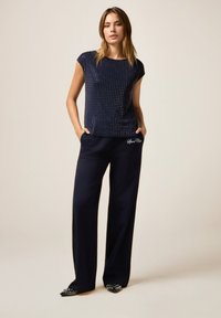 Marineblauer Kurzarm-Oberteil mit silbernen Punkten, kombiniert mit marineblauen weiten Hosen mit weiß gesticktem Text; schwarze spitze Schuhe mit Nieten.