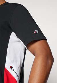 Camiseta de algodón negra con paneles laterales en blanco y rojo, un logo de Champion en la manga y un diseño ajustado con una textura suave.