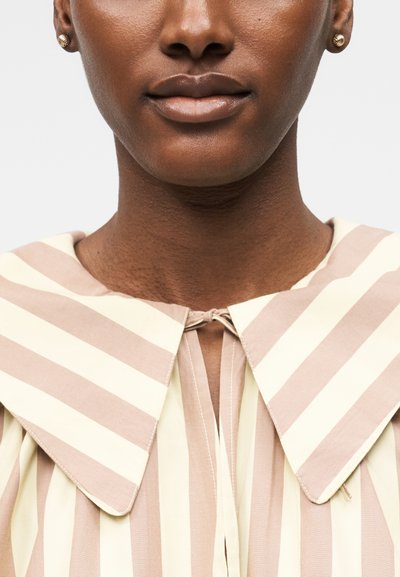 Κοντινό πλάνο ενός ατόμου που φοράει μπλούζα με μπεζ και λευκές ρίγες, με υπερμεγέθη μυτερή γιακά και χρυσά σπειροειδή σκουλαρίκια.