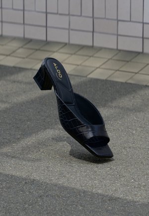 Sandalia mule negra con tacón con estampado de cocodrilo, punta cuadrada y tacón ancho, descansando sobre suelo texturizado cerca de una pared con azulejos.