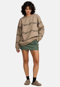 Felpa beige con design floreale nero, abbinata a una minigonna verde. L'outfit presenta una vestibilità comoda e tessuti morbidi.